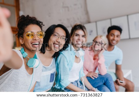 Laughing african girl in headphones and yellow glasses making selfie sitting on the bench with university mates. Excited female student taking picture of herself and her asian friend with long hair.