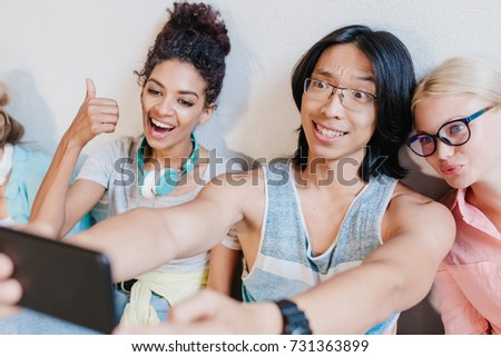 Asian long-haired boy making selfie with friends holding black phone and smiling. Funny blonde girl in glasses having fun with curly young lady and brunette student.