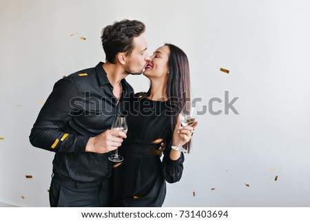 Black-haired man in stylish shirt kissing his wife on light background. Cute couple with glasses of wine celebrating anniversary under sparkle confetti.