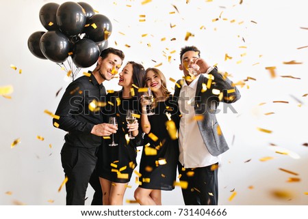 Dreamy blonde girl with glass in hand looking at confetti while her african friend drinking wine. Indoor portrait of young people chilling at party with balloons.