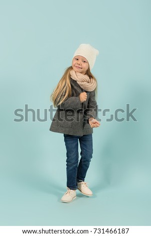 Vertical picture, isolated on blue, beautiful caucasian blonde little girl in grey winter coat, jeans, white hat and scarf stands