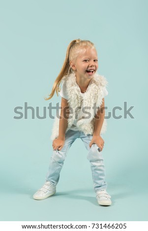 Vertical picture, isolated on blue, pretty caucasian blonde little girl in white t-shirt, fur jacket, light blue trousers and white shoes stands bended, laughing