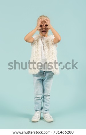 Vertical picture, isolated on blue, pretty caucasian blonde little girl in white t-shirt, fur jacket, light blue trousers and white shoes stands, hands on the face