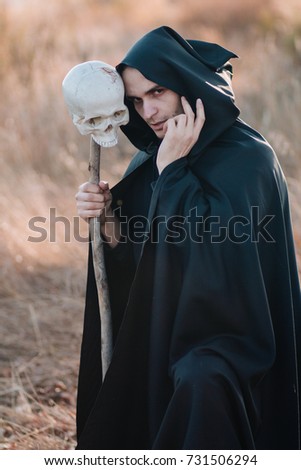 Halloween image. A handsome guy in a black cloak with a staff from a human skull against the background of a yellow field. Fantasy
