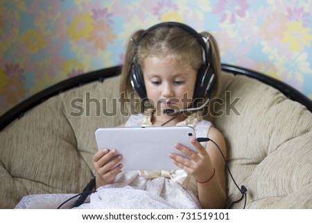 little girl in a beautiful dress in headphones with a microphone sitting in a chair and looks at the tablet