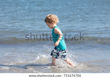 A little boy with blonde curly hair is splashing and playing at the beach. He is having fun in the waves on a sunny day. 