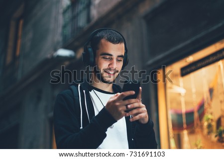 Handsome young man in black hoodie wears big wireless music headphones and changes audio tracks on smartphone, he smiles when looks at phone screen Royalty-Free Stock Photo #731691103