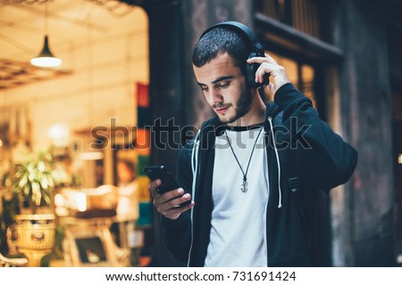 Hispanic young attractive man stands in dark street in front of shop, changes songs and tracks on smartphone, listens to music in wireless headphones. Hipster with slight beard Royalty-Free Stock Photo #731691424