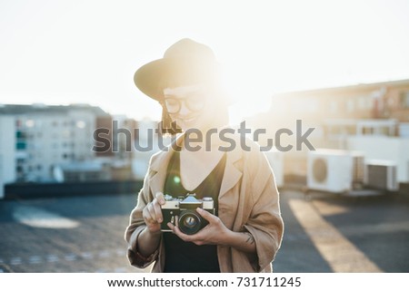 Beautiful young woman stands on top of rooftop in middle of big city with amazing shadows and sun beams and leaks behind her, holds vintage analog camera