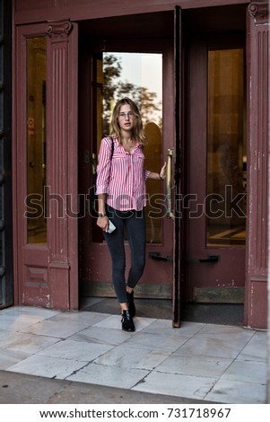 Pretty and cute feminine woman in skinny jeans and red shirt leaves building through vintage wooden rotating doors on warm summer evening, on way to business meeting 