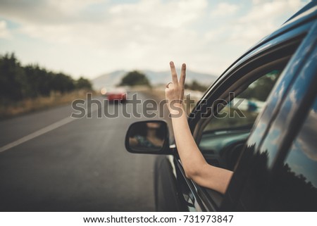 Young happy young girl drives a car a holds her hand out from the window. Lady driver enjoys driving and shows Ok sign with her hand out of window. Road trip, travel and freedom concept.