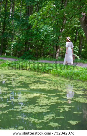 beautiful woman in a white dress and hat goes along the shore of a pond in the park