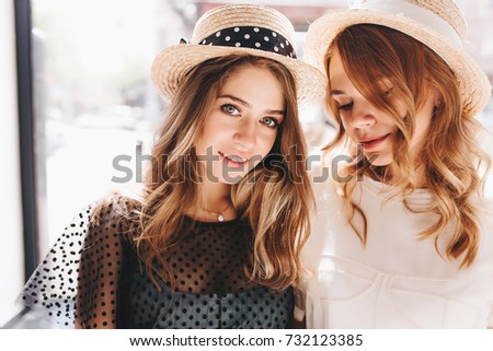 Close-up portrait of girl with gray eyes wears straw hat with ribbon gladly posing beside curly sister. Two fascinating ladies in elegant attire spending time together enjoying weekend.