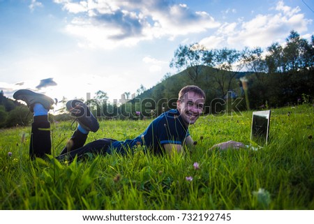Young man lies on green grass at sunset with laptop and working 