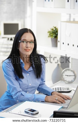 Portrait of smiling assistant girl wearing glasses, sitting at office desk with laptop computer, looking at camera.?