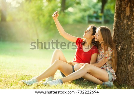 Two Beautiful Teenagers Make Selfies, Sitting under a Tree in the Bright Summer Day
