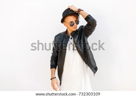 Fashionable handsome man in a black stylish coat and hat with sunglasses posing near white metal wall