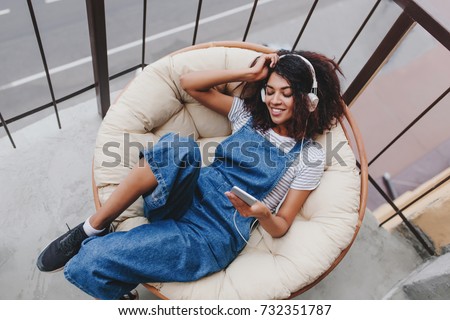 Pleased black girl wearing trendy sport shoes chilling on chair on balcony, enjoying morning alone. Portrait of smiling cute young woman in denim clothes having fun on terrace in weekend. Royalty-Free Stock Photo #732351787