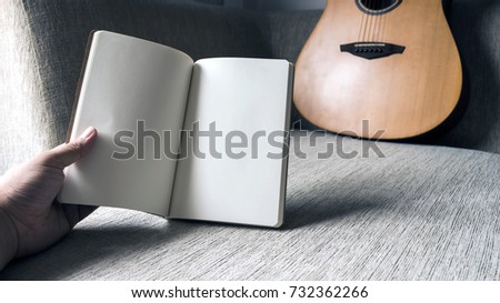 Holding a blank notebook on the sofa, which has a guitar and soft light through from the outside. Pallid color picture tone 