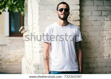 Young stylish man wearing white blank design t-shirt with beard in glasses, standing on the street on city background. Street photo
