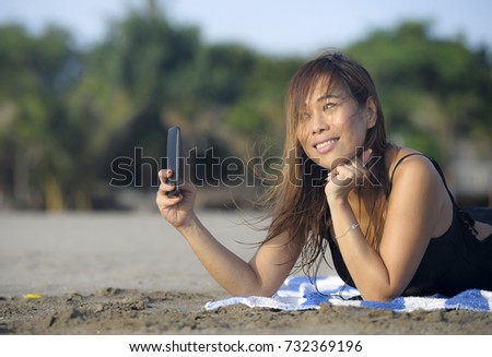 young beautiful and happy Asian woman using mobile phone taking selfie portrait photo having fun relaxed on beach sand having suntan enjoying tropical holiday and vacation