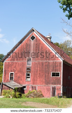 Rustic old red barn in disrepair with grass in the foreground and blue sky above.  A weathervane sits on the roof of the barn.