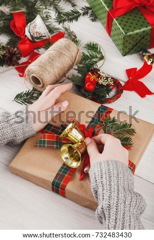 Creative diy hobby. Decorating modern handmade xmas present with bells. Box in craft paper with checkered ribbon. Woman's hands on white wood table with fir tree branches, decoration of gift.