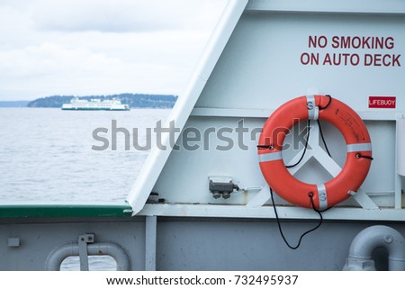 Lifebuoy ring hanging on the port hull of an industrial ship, with a sailing ferry boat and coastline in the background
