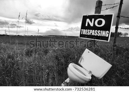 A black and white image of a no trespassing sign and old toilet in a field. 