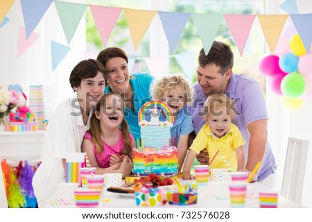 Kids birthday party with unicorn rainbow cake for little girl. Family celebrating child birthday. Parents and children blowing candles at baby cake. Balloons and colorful pastel banner decoration.