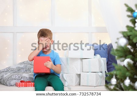 Boy in a blue t-shirt opens a box with a Christmas present sitting on a window sill next to a Christmas tree