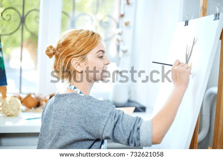 Young smiling girl paints on canvas with oil colors in own workshop. Window on the background. Art concept.
