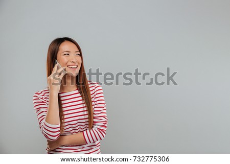 Picture of laughing young asian woman talking by phone standing isolated over grey wall. Looking aside.