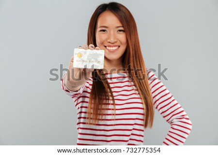 Picture of young asian happy lady standing isolated over grey wall. Looking camera holding credit card. Focus on card.
