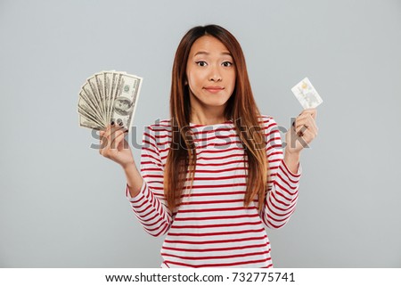 Picture of pretty young asian lady standing isolated over grey wall. Looking camera holding money and credit card.
