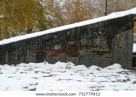 An old, shattered gray and black wall with red brick, green moss, white snow, snowdrifts. Slanting roof, diagonally. Trees with yellow leaves. Autumn and winter background