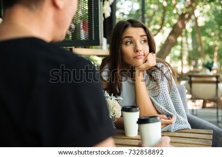 Tired bored girl sitting and drinking coffee with her boyfriend at a cafe outdoors and looking away