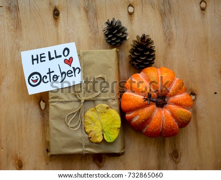 Rustic gift in paper craft with Hello October text on a note and a pumpkin ,pine cones and autumn leaf on wooden table