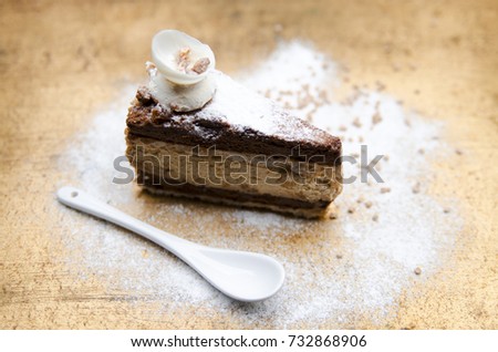 Piece of creamy cake with vanilla and cocoa cream. Slice of tasty pie arranged with sugar powder and small white spoon on golden rustic table.