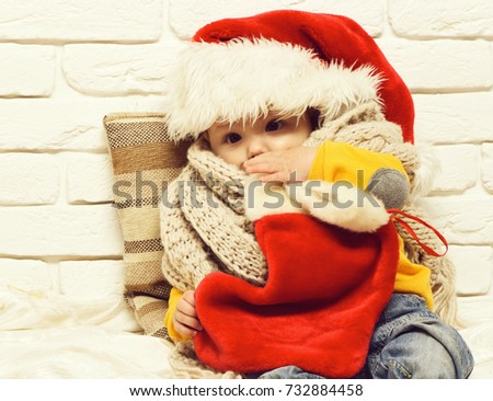 small baby boy with adorable curious face in red sweater with dummy in new year hat scarf and christmas or xmas stocking or boot on white brick wall background