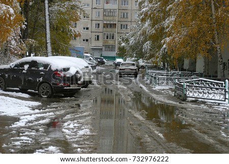 Wet road, sidewalk, iron black decorative fence covered with white fluffy snow, parked cars in the city yard. The first snow in autumn, in October. Trees with yellow leaves. Winter background