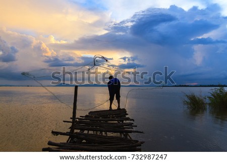 Thai fisherman on a wooden bridge casting a net for catching freshwater fish in nature river in the early evening before sunset