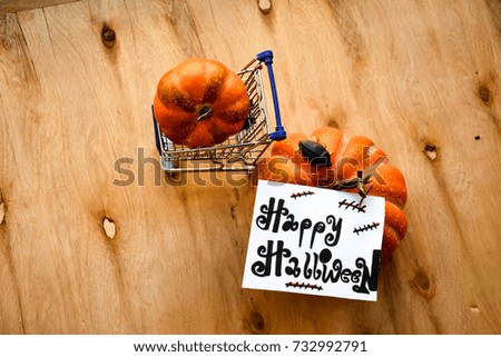 A shopping cart with a pumpkin inside and a pumpkin near and Happy Halloween text on a note on wooden table