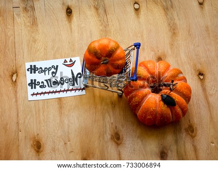 A shopping cart with a pumpkin inside and a pumpkin near and Happy Halloween text on a note on wooden table