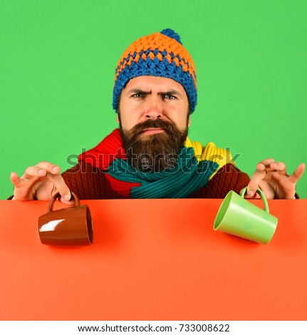 October beverage idea. Hipster with beard and confused face has warm tea or coffee. Autumn and cold weather concept. Man in hat holds brown and green cup on green and orange background, copy space