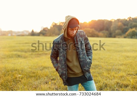 Handsome man with a cap in a military jacket with a hoodie on the nature at sunset