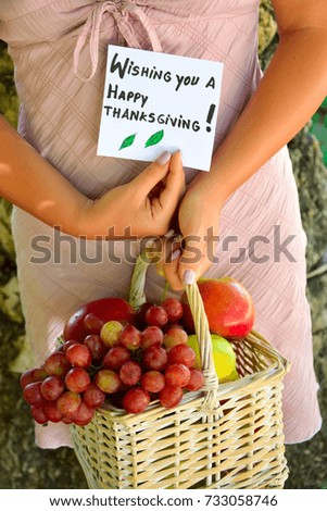 Woman holding a basket of fresh fruits and wishing you a Happy Thanksgiving text on a note in a green garden