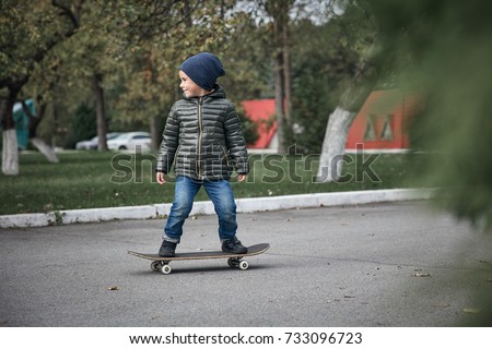 Cute little boy wearing dark grey jacket, dark blue cap and blue jeans, riding on grey skate board outdoors  