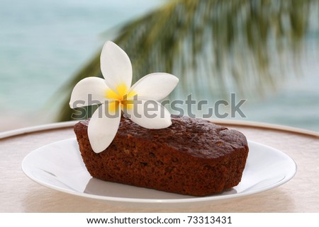 traditional hawaiian banana bread with a hot pink plumeria flower on a white plate outside with the pacific ocean out of focus in the background on the island of maui hawaii