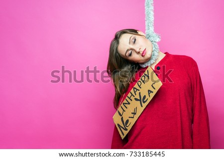 A beautiful overwhelmed woman in red pajamas with a sign "unhappy new year" hanged herself on a Christmas garland from the festive stress, on a pink background. Vanity, holiday problems, rush